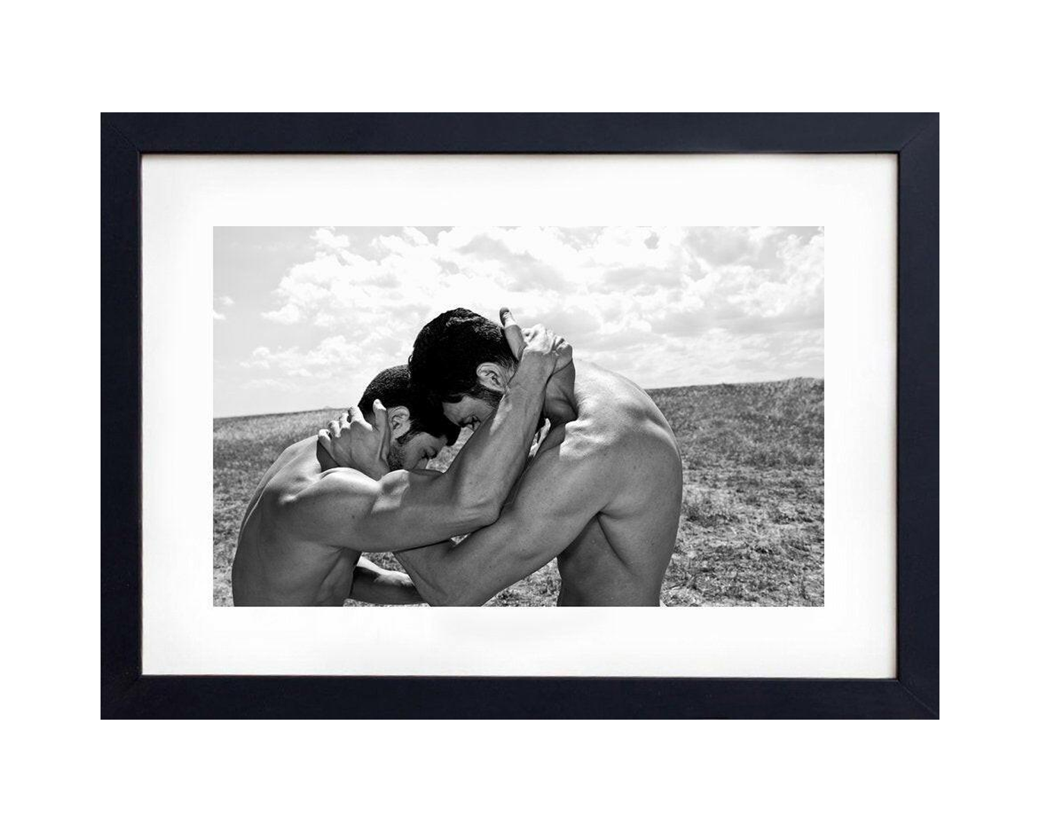 Framed black and white photograph of Two men engage in a dynamic embrace in a barren landscape, showcasing strength and emotion against a cloudy sky in black and white.