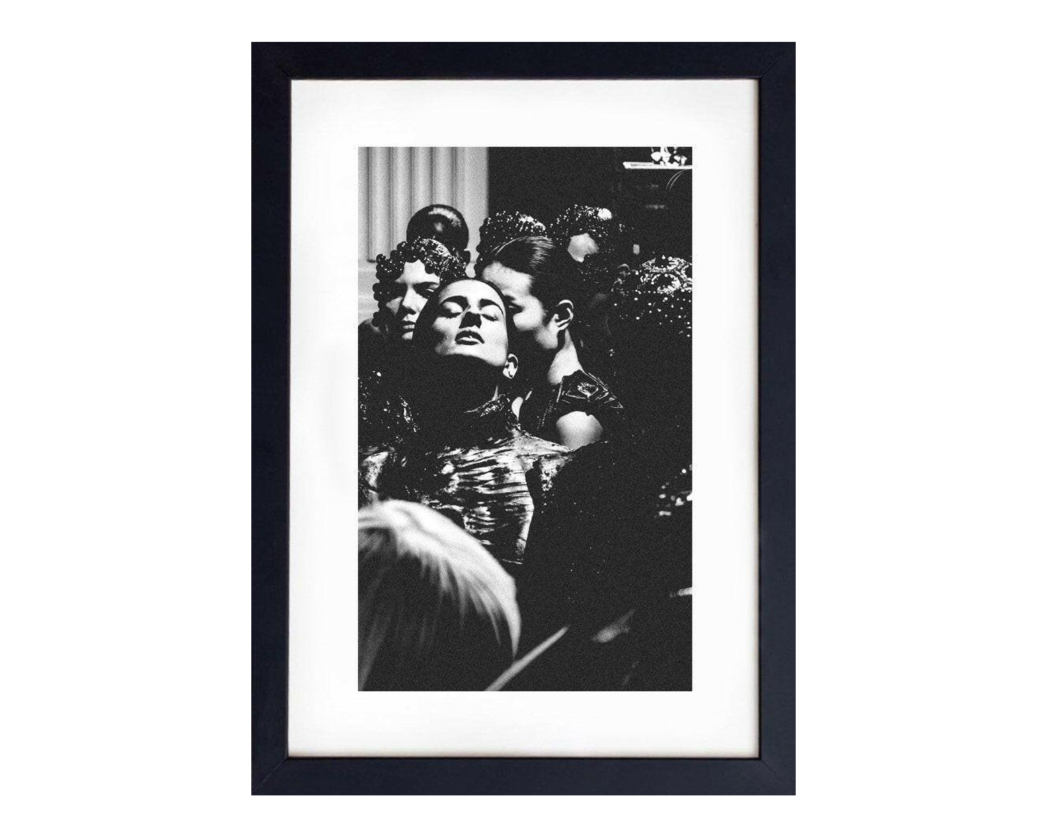 Framed black and white image of models backstage, featuring intricate hairstyles and adorned outfits, preparing for a fashion show.