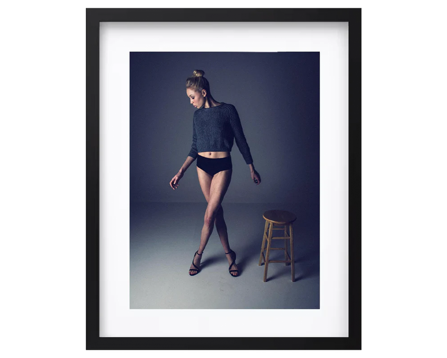 Framed photograph of a woman in a dark outfit standing next to a wooden stool against a dark background.