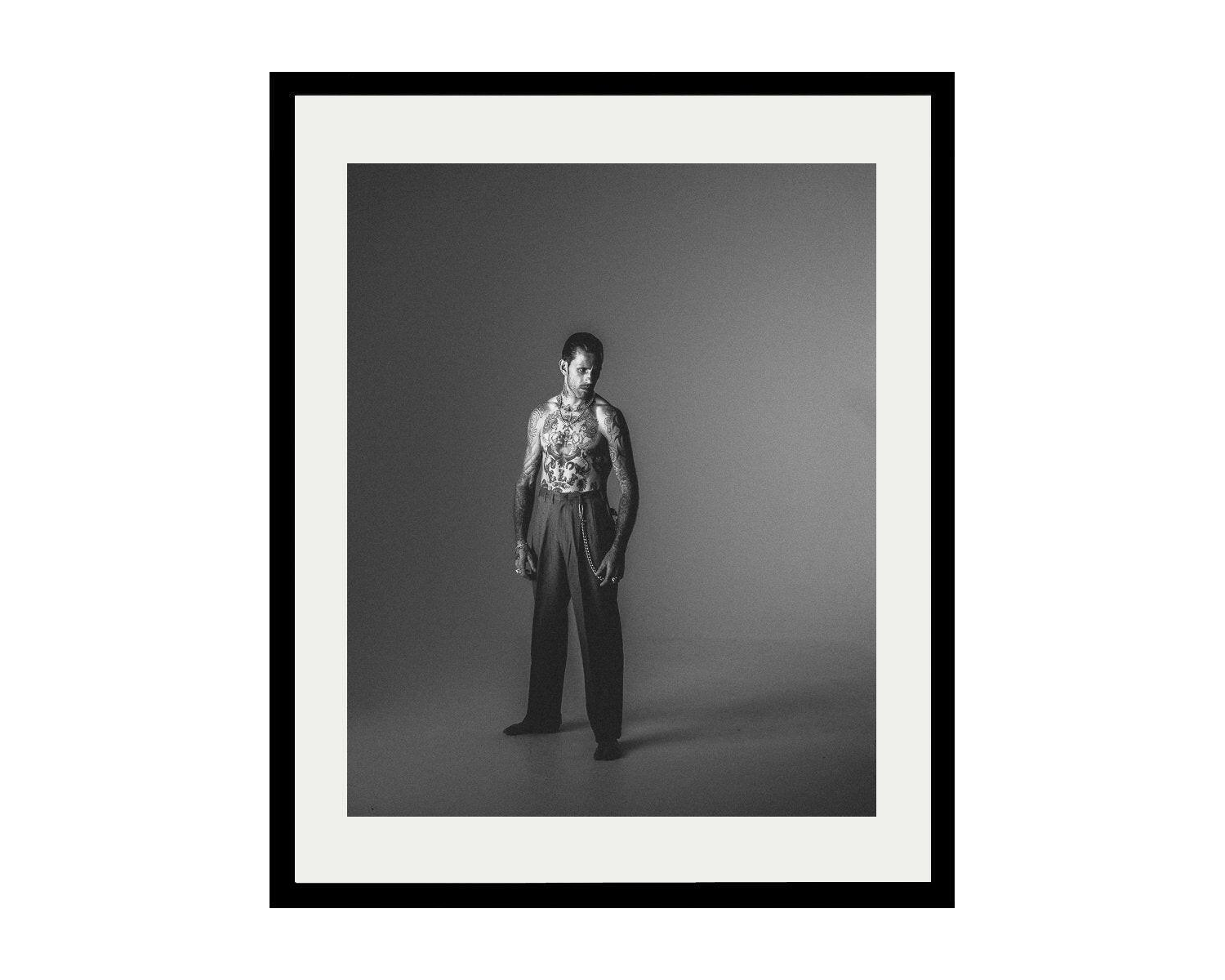 Framed black and white portrait of A tattooed man  positioned in a shadowy room, wearing a pant, with the darkness adding to the scene's intrigue
