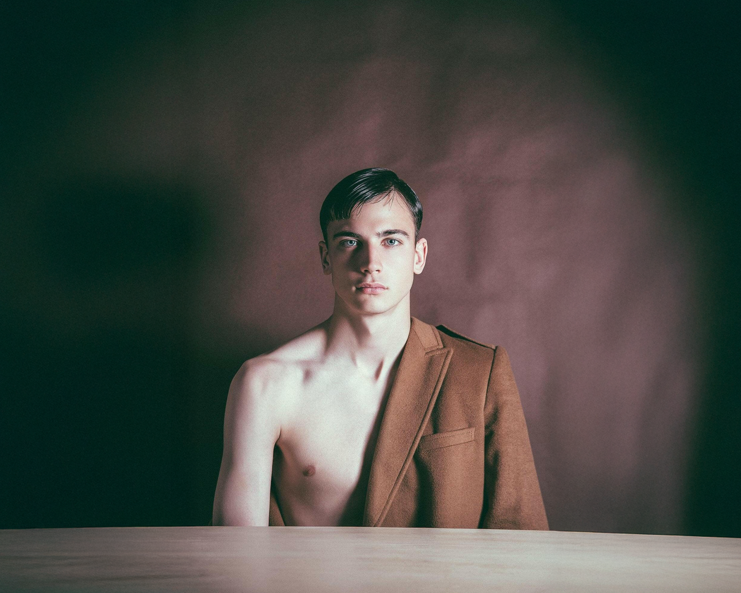 Man wearing a brown jacket without a shirt, sitting at a table with a dark background