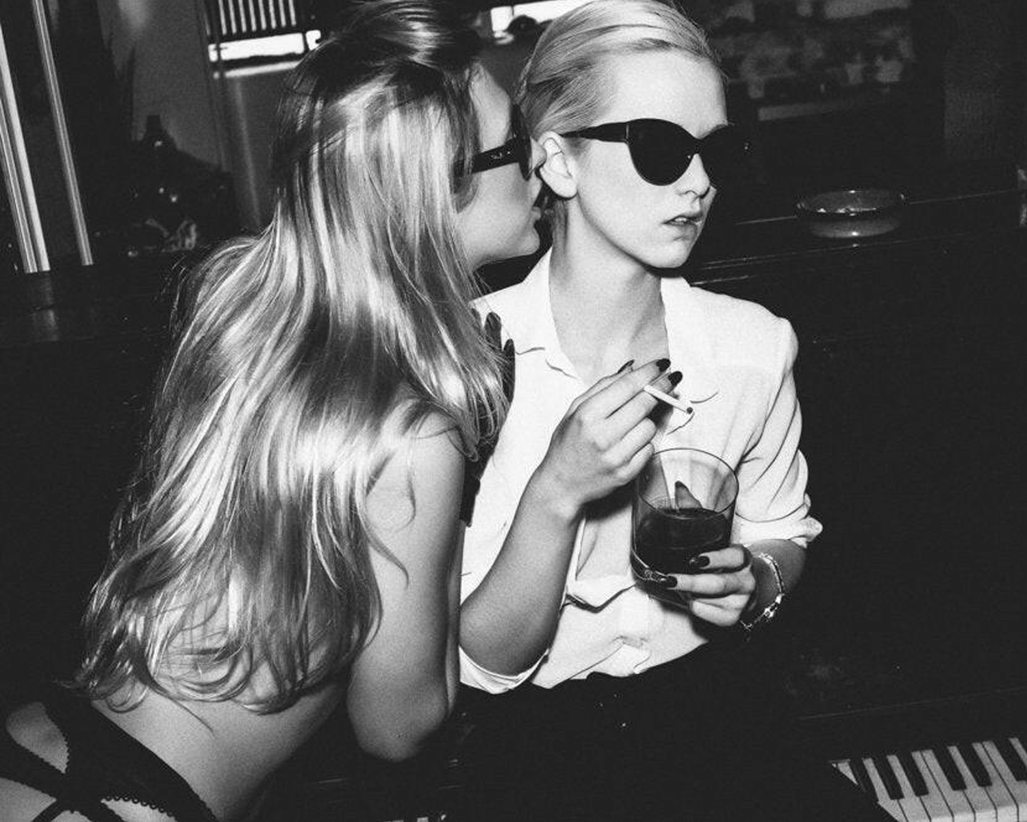 Two women in a social setting, one whispering to the other and holding a glass of red wine.