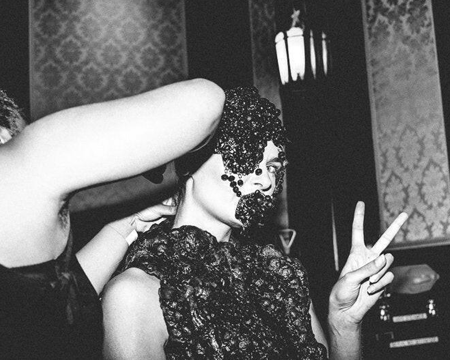 Person wearing a textured mask and black lace dress in a room with patterned wallpaper.