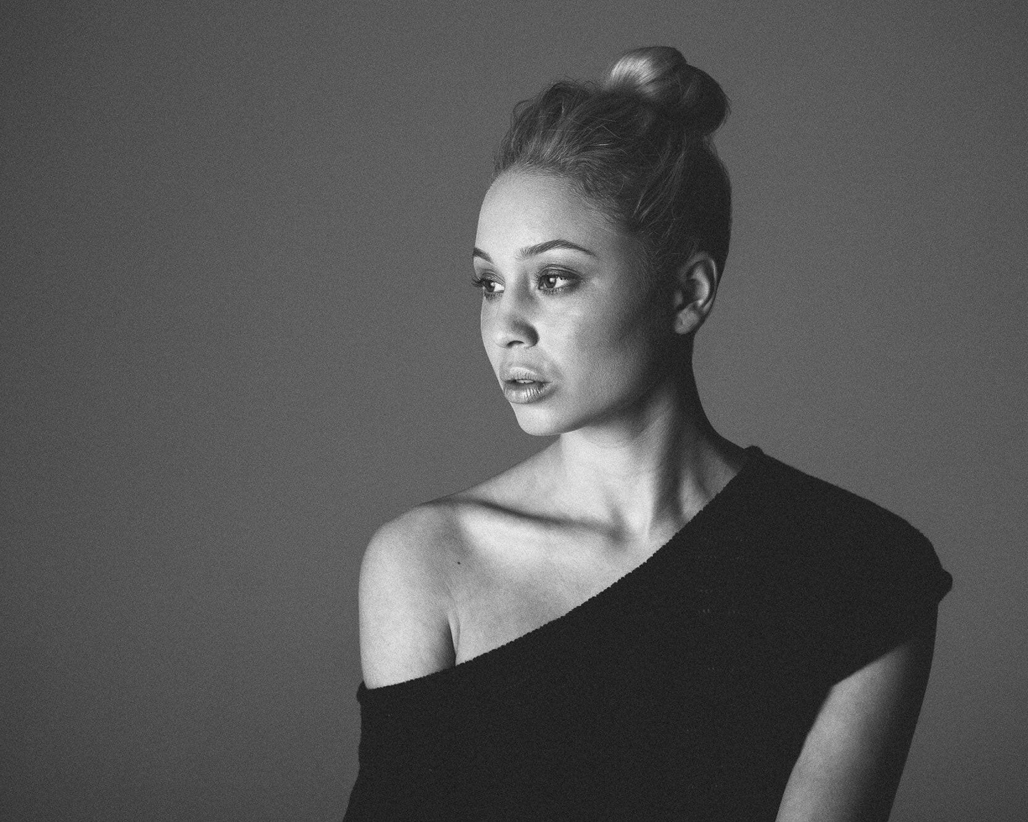 Black and white portrait of a woman with a bun wearing a one-shoulder dress against a gray background