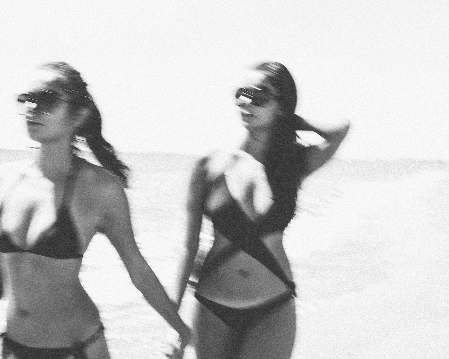 Two women in black swimsuits walking on a beach.