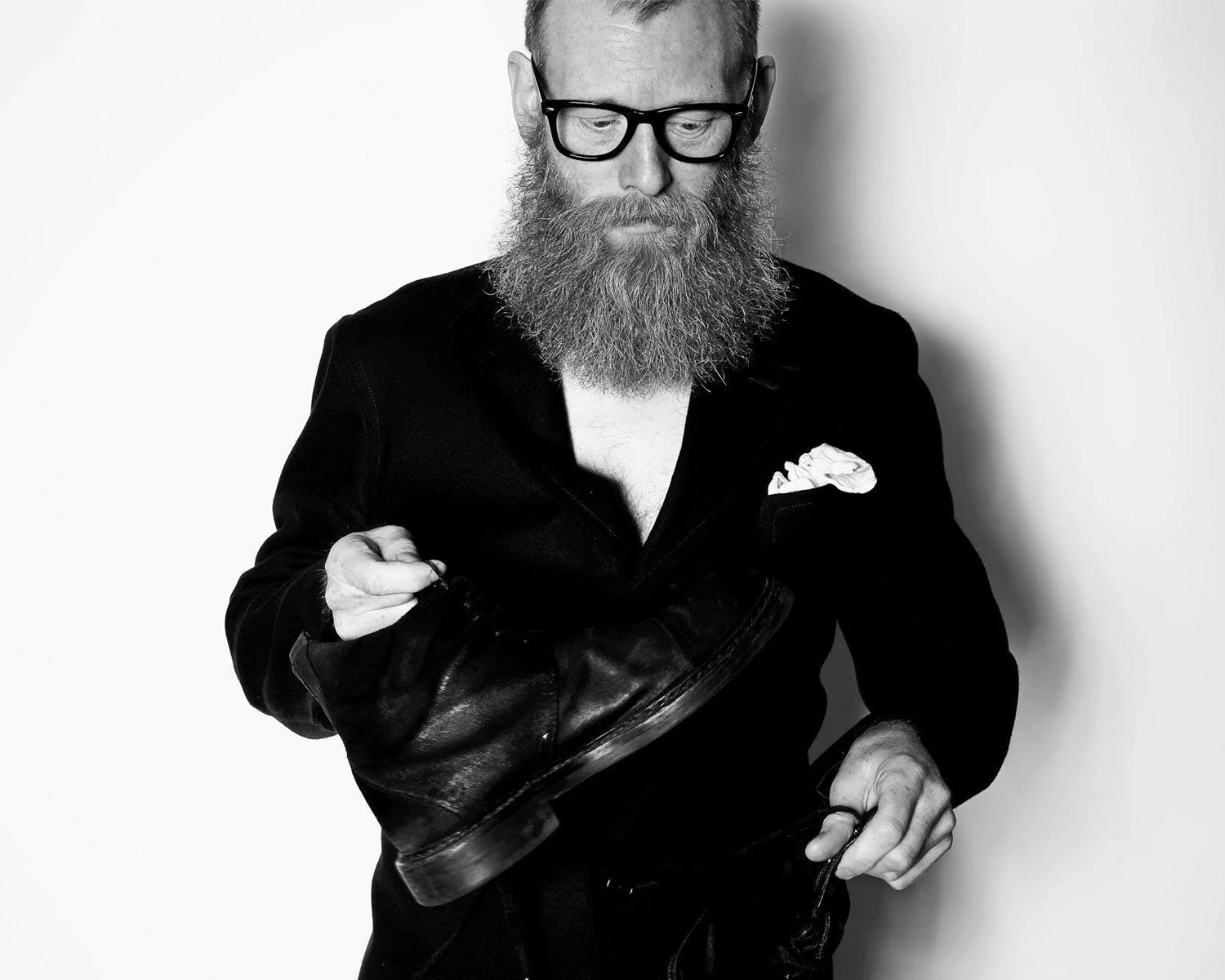 Man with a beard wearing glasses and a black jacket holding a leather hat against a white background