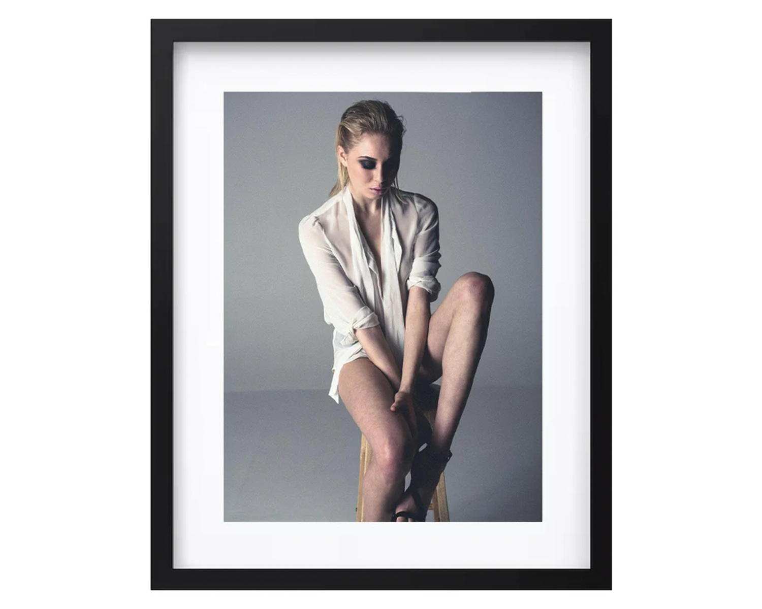 A woman in a white shirt and black shoes is seated on a stool, with a relaxed posture and a neutral expression, framed in black.